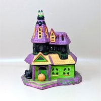 A vintage Celebrations by Silvestri ceramic Halloween house. It features a pointed purple roof, a bright green facade, and playful details like an owl on the tower, bats, and a small jack-o'-lantern at the front entrance.