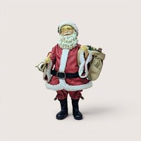 Standing Santa Claus figurine with glasses holding a gold handbell and carrying a sack of toys over his shoulder, centered on a plain linen background.