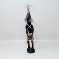 A vintage hand-carved wooden African tribal figure. The sculpture features dark stained wood, painted details in orange on the shorts and headdress, and decorative white fiber straps with genuine feather embellishments.