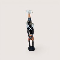 Carved wooden tribal figurine standing on a circular base, featuring feather details on the headpiece and shoulders with a white criss-cross chest pattern, centered on a plain linen background.