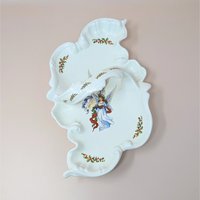 A vintage handmade ceramic serving tray with a sculpted, flowing edge. The white glaze is decorated with hand-painted holly sprigs and a central illustration of an angel, with a large integrated handle.