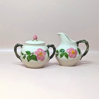 Hand painted Franciscan Desert Rose ceramic cream and sugar set on a soft pink background. The lidded sugar bowl and cream pitcher feature dimensional pink wild roses with green leaves and realistic brown branch handles and finial.