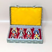 A vintage boxed display of eight hand painted Beijing Opera clay masks resting on a soft pink background. The vibrant, colorful theatrical facial makeup designs are arranged on red velvet inside a fabric covered case with a bright yellow satin lined lid.