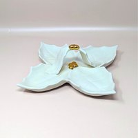 A vintage cream colored Holland Mold ceramic four part holly leaf dish resting on a soft pink background. The square hostess tray features a prominent central arched handle and hand painted shiny gold metallic berry clusters at the center and apex.