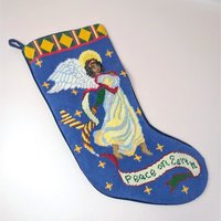A vintage handmade needlepoint Christmas stocking on a soft pink background. The blue stocking features a hand stitched Black angel with white wings and a gold halo, surrounded by yellow stars and holding a banner reading Peace on Earth.