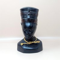A vintage hand carved wooden bust of Egyptian Queen Nefertiti resting on a soft pink background. The dark polished sculpture features a tall flat topped crown with carved bands and a simple tan fiber rope knotted around the neck.