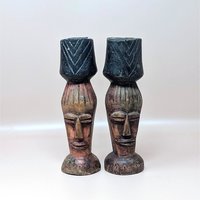 A pair of vintage hand carved wooden candlestick holders on a soft pink background. The dark brown pillars feature stylized tribal or tiki faces topped with dark, chevron carved candle cups and flare out into circular bases.