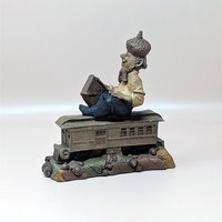 A vintage 1983 Tom Clark resin gnome figurine named Rolf on a soft pink background. The highly detailed statue shows a smiling bearded gnome wearing an acorn hat sitting on top of a brown railway car with a suitcase.