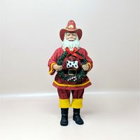 A vintage Fabriche fireman Santa Claus figurine on a soft pink background. Standing eleven and a half inches tall, the figure wears a red firefighter uniform and hat while holding a green faux pine wreath adorned with a spotted dog bone and red ribbon.