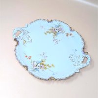 A vintage Rosenthal porcelain handled cake plate resting on a soft pink background. The antique German serving tray features delicate pink and yellow floral bouquets, an ornate scalloped edge, and brushed gold accents on the handles.