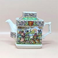 A vintage Sadler porcelain castle themed teapot on a soft pink background. The square cream colored teapot features a detailed double sided illustration of the Sheriff of Nottingham at his table and Robin Hood in Sherwood Forest with stone walls.