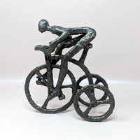 A vintage brutalist cast iron sculpture of a man riding a tricycle on a soft pink background. The dark, heavily textured metal figurine depicts a stylized, faceless human figure leaning forward in a racing position over three spoked wheels.