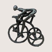 A vintage brutalist cast iron sculpture of a man riding a tricycle on a soft pink background. The dark, heavily textured metal figurine depicts a stylized, faceless human figure leaning forward in a racing position over three spoked wheels.