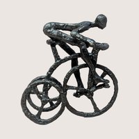A vintage brutalist cast iron sculpture of a man riding a tricycle on a soft pink background. The dark, heavily textured metal figurine depicts a stylized, faceless human figure leaning forward in a racing position over three spoked wheels.