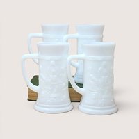 Four vintage white milk glass mugs with raised tavern scenes arranged around a green book on a plain background