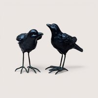 Two black ceramic crow figurines with long metal legs standing side by side on a plain background