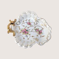 A vintage white porcelain shell dish with pink rose bouquets and an ornate gold handle on a plain cream background