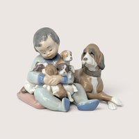 A vintage porcelain figurine of a young boy sitting on a pink cushion holding four puppies next to a large dog