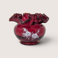 A vintage ruby red glass vase with a deeply ruffled rim and hand painted white roses on a plain background