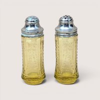 A pair of amber depression glass salt and pepper shakers with silver metal lids on a plain beige background