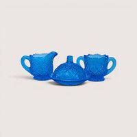 A miniature blue glass toy creamer, lidded butter dish, and open sugar bowl set on a plain cream background