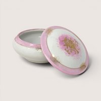 Round white porcelain trinket box with an open lid featuring a pink rim and a gold floral basket on a solid background.