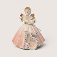 Porcelain birthday angel figurine in a pink dress holding a gift behind a floral number 15 on a solid background.
