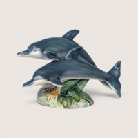 Two glossy blue-grey ceramic dolphins leaping together over a green sea coral base on a solid background.