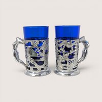 Pair of vintage silver plated metal dragon wrap tankards with cobalt blue glass inserts on a solid background.
