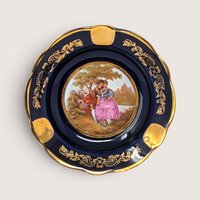 A round cobalt blue porcelain ashtray with gold scrolling trim and a central romantic couple scene on a white background