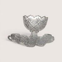An antique clear pressed glass miniature punch bowl on a pedestal with six matching tiny cups on a plain background