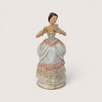 An antique porcelain figurine of a lady holding a hand fan with pink lace style details on a plain background