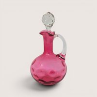 Cranberry glass cruet bottle with a clear applied handle and a faceted clear glass stopper against a neutral background