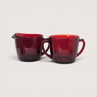 A vintage deep red glass creamer pitcher and matching two handled sugar bowl side by side on a plain background