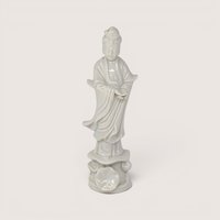 A vintage white blanc de chine porcelain Guan Yin figurine standing on a lotus base against a plain studio backdrop