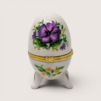 Vintage porcelain egg-shaped trinket box with a painted purple flower and gold feet set against a plain linen background.