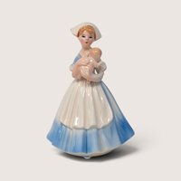 Vintage porcelain figurine of a nurse in a blue and white dress holding a baby, with a close-up highlighting a small chip on her hat.