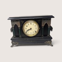 Front view of the antique black architectural mantel clock showing the detailed dial with arabic numerals, ornate side pillars, and decorative cast metal feet.   