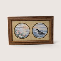 A vintage framed print by D. Smith featuring two circular windows set in a textured fabric mat: one showing white lady's slipper wildflowers and the other a loon swimming on a calm lake, enclosed in a wooden frame.