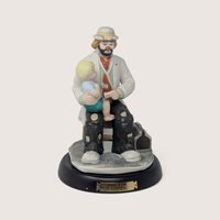 A vintage Flambro Emmett Kelly Jr. porcelain doctor figurine featuring a clown holding a child on a wooden base.