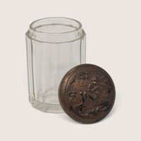 A vintage faceted glass jar with an ornate embossed metal lid showing a seated figure.