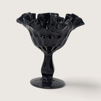 A vintage Fenton black glass compote with an optic thumbprint pattern and a ruffled rim on a neutral background.