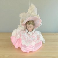 A decorative Christmas ornament featuring a doll head with closed eyes and blonde hair. She is wearing an oversized pink and white lace sun hat and a matching voluminous pink satin dress with a tiered white lace collar and overlay.