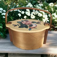 A vintage wooden woven basket with a hinged lid featuring a hand-painted floral design of a pink rose and blue flowers, with a stationary arched wood handle.