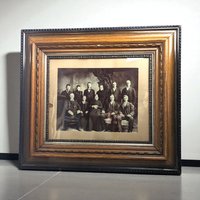 Antique black and white family portrait in a wide, ornate wooden frame. The frame features deep wood grain, silver-toned inner borders, and decorative beadwork. Sepia-toned image shows eleven family members in formal period attire.