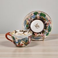 A white Italian porcelain teacup and saucer set featuring highly detailed, hand-painted raised relief figures of classical men and women in a nature landscape with elegant gold accents.