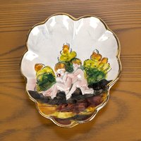 A white heart-shaped or oval ceramic trinket dish featuring hand-painted raised relief figures of children playing in a nature scene with a gold-gilded rim.