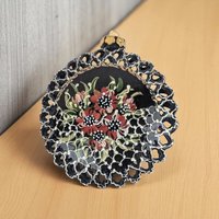 A circular, flat-backed glass ornament featuring a black base with hand-painted red and white floral details. The perimeter is defined by an intricate, die-cut lattice border, while the center displays a bouquet of flowers with white dot accents.