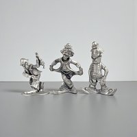 Three small solid pewter clown figurines in various playful poses. The metal has a cool grey, matte patina with dark oxidation in the recessed details. Their silhouettes are slim and whimsical, featuring oversized shoes and tiny hats.