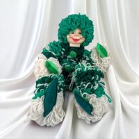 A sitting clown with a smiling plastic face and a body made entirely of looped green and white yarn. It features dark green felt hands and feet. The yarn creates a thick, curly texture over the entire silhouette.